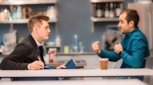 Two students sit at a table together. One is holding a notebook and listening while the other student gestures with their hands. They appear to be sitting at a coffee stop or restaurant on a CFE trek.