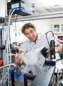 A man smiles for the camera while pouring a smoking liquid into another container in a lab setting