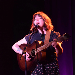 Woman with brown hair singing into microphone and playing guitar