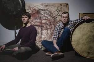 2 men sitting in front of abstract painting and next to drum