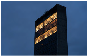 A black building, windows lit from within, is seen from below against a solid blue sky