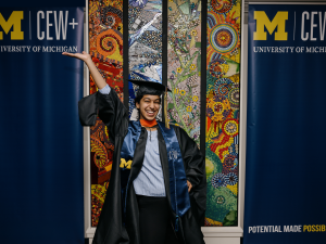 A joyful graduate in University of Michigan cap and gown poses with one arm raised, set against a vibrant mosaic backdrop. Two university banners flank her.
