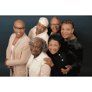 Portrait of six Black women standing closely together against a dark blue background. Some have bald or closely cropped hair, while others wear short natural styles or a headscarf. They are smiling and leaning into one another with arms around each other.