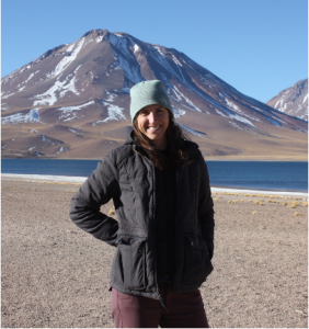 Person wearing a light blue knit beanie and black puffer jacket stands on a rocky shoreline beside a blue lake, with a snow-streaked mountain rising in the background under a clear sky.