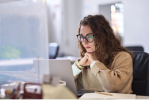 Person with curly hair and glasses sits in a chair at a desk, hands under their chin, looking at a laptop in a bright office.