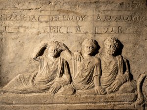 Close-up of a Roman Egyptian stone stela with a carving of three people wearing draped clothing reclining on a couch, with Greek inscriptions above each of their heads.
