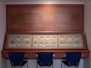 Section at the Kelsey Museum that teaches about seals and sealings in ancient Mesopotamia. The display features a long case with three chairs in front of it. The case contains some two dozen ancient cylinder/rectangular seals, modern impressions of each object in clay, and small paper labels showing illustrations of the full designs at a larger size.