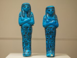 Two bright-blue small faience ushabtis stand on display side by side with their arms crossed over their chests. Black paint defines their facial features and hair, while several registers of hieroglyphs appear on their legs.