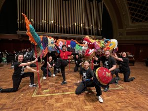 A group of dragon dancers wearing all black are lined up on a stage holding up a dragon sculpture made out of colorful fabrics