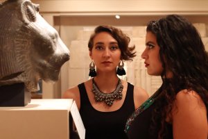 Two women in a museum gallery space; Nabra Nelson stares directly at the camera, while Sarah Fahmy, in profile, views a stone sculpture of a lion’s head.