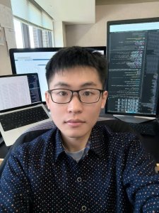 A man in glasses and a navy shirt sits in front of several computer monitors