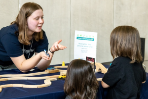 A young woman leans over a table and speaks to two children