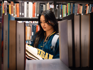 A woman wearing a Michigan sweatshirt reads a book in a library aisle, surrounded by shelves filled with books.