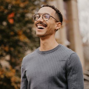A young man with glasses and a mustache laughing outdoors against a blurred autumn background.