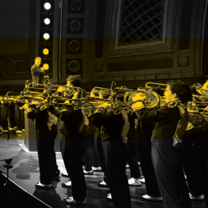 Michigan Marching Band members play brass instruments onstage while a conductor leads them under bright stage lights.