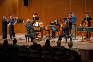 A small ensemble of strings and early keyboards performs on stage, with an audience seated in the lower foreground.