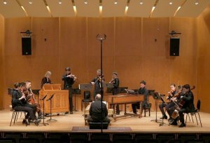 A small ensemble of strings and early keyboards performs on stage, with an audience seated in the lower foreground.