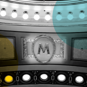 Ornate “M” emblem centered over a row of bright white stage lights, accented by soft yellow and blue circles.