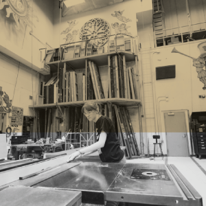 Person working with a table saw in a creative, organized workshop with large painted murals and stored materials.