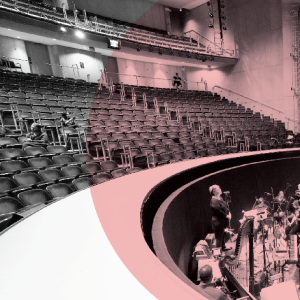 Spacious theater within the Power Center with rows of empty seats, only a handful of people scattered throughout. Musicians and a conductor gather in the orchestra pit, ready for rehearsal.