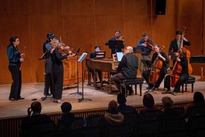 A small ensemble of strings, early keyboards, and a wind player performs on stage, with an audience seated in the lower foreground.