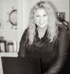 Black-and-white portrait of a smiling woman with long curly hair seated at a table with a laptop.