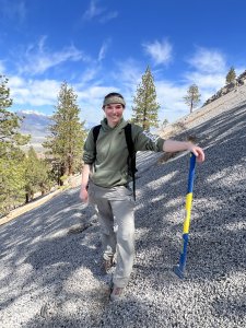 A woman in khakis and a green sweatshirt smiles at the camera from the side of a gravelly mountain