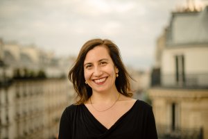 A woman with shoulder-length brown hair, a black top, and subtle gold jewelry smiles at the camera. She is positioned against a softly blurred background of architectural rooftops under an overcast sky.