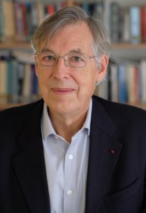 A headshot of Francois Heisbourg wearing a dark suit jacket over a light blue collared shirt and thin-framed glasses. He has grey hair parted to the side and is smiling faintly against a blurred background of bookshelves.