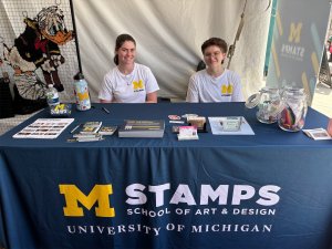 Two students, smiling, sit at a table covered with a blue table adorned with an M STAMPS logo