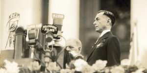 A man in a suit - Manuel Quezon - standing near a bank of microphones at an outdoor speech.