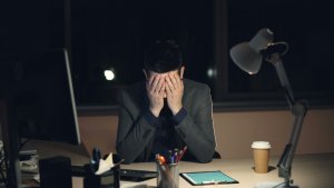 Tired young man in suit and tie is working on computer in office at night sitting at desk in dark room, looking at screen and typing. Hard work, youth and stress concept.