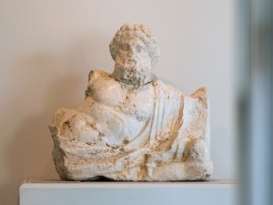 White travertine sculpture of the torso and head of Nilus, a bearded and curly-haired male figure in a semi-reclined pose.
