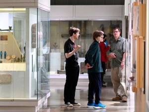 A family of visitors pauses between the Dynastic and Graeco-Roman Egyptian galleries at the Kelsey Museum. Displayed in a case to their left are several small Egyptian artifacts, including bronze figurines of deities and faience ushabtis.