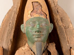 Close-up of a figurine of the Egyptian god Osiris, with a green-painted face, a long goatee, and an Atef crown, housed within a small wooden coffin.