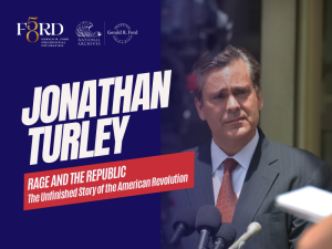 A photo of Jonathan Turley wearing a grey suit with a red paisley tie standing at a podium