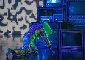 A dancer crawls near a grouping of televisions, in futuristic silvery costume.