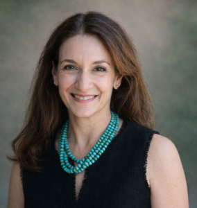 Portrait of Stacey Rudnick smiling at the camera against a softly blurred gray-green background. She has long, dark brown hair worn down and is dressed in a sleeveless black top with a layered turquoise beaded necklace and small stud earrings. The portrait is framed from the chest up with natural lighting.