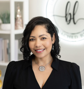 Portrait of Dr. Barbara Lash, ’92, smiling at the camera in a bright office setting. She has shoulder-length black wavy hair and is wearing a black V-neck blouse, silver drop earrings, and a silver pendant necklace with a circular floral design. Shelving and a blurred business logo are visible in the background, and the portrait is framed from the chest up with soft, natural lighting.