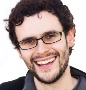 Portrait of Jeremy Schifeling, MBA ’12, smiling broadly at the camera against a plain white background. He has short, curly dark hair, wears black rectangular glasses, and has a neatly trimmed beard and mustache. He is dressed in a dark zip-up jacket over a gray shirt. The portrait is closely cropped around his head and shoulders with bright, even lighting.