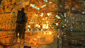 An artist stands on a wooden ladder to paint a massive, warm-toned abstract mural covered in calligraphic symbols and dense, dripping paint textures.