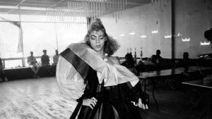 A black-and-white photograph of Octavia St. Laurent posing dramatically in an elaborate, voluminous gown inside a hall with trophies and onlookers in the background.