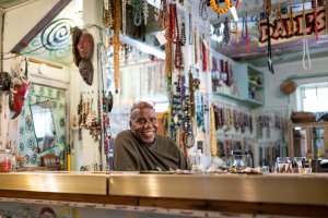 Olayami Dabls smiling behind a counter inside the museum filled with hanging beads, masks, and artwork.