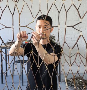 Artist Tomoyuki Ueno carefully uses pliers to attach small wooden twigs together with metal jump rings, constructing a large hanging screen.