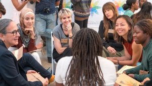 A diverse group of people seated in a circle engaging in a story exchange discussion, holding folders and listening intently to a speaker in the foreground.