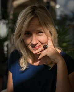 A blonde woman wearing a dark blue shirt smiles with her hand resting against her chin, wearing a large dark ring.