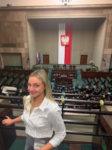 A blonde woman wearing a white collared shirt stands on a balcony overlooking a large legislative assembly hall.