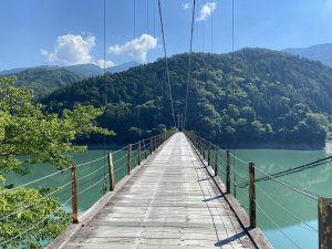 A photo of a wooden footbridge over a wide river in the Japanese mountains on a clear day.