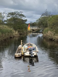 A small motorized boat with a small wooden vessel latched to the side, floating on a small river with a small concrete bridge in the background.