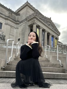 A photo of the speaker, Kanako Uzawa. She is a crouching outside of a large gray museum-like building, looking pensive up at the sky.